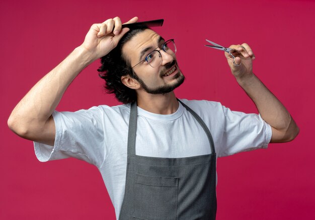 Impressed young caucasian male barber wearing glasses and wavy hair band in uniform combing his hair and holding scissors looking at side isolated on crimson background