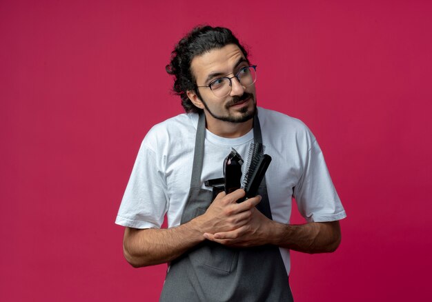 Impressed young caucasian male barber wearing glasses and wavy hair band in uniform holding barber tools looking at side isolated on crimson background with copy space