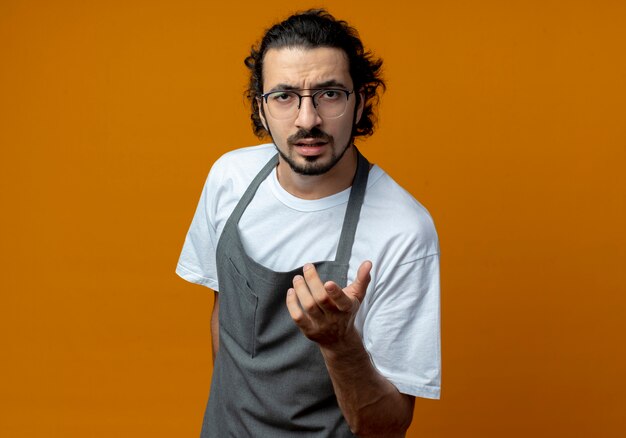 Impressed young caucasian male barber wearing uniform and glasses pointing at camera isolated on orange background with copy space