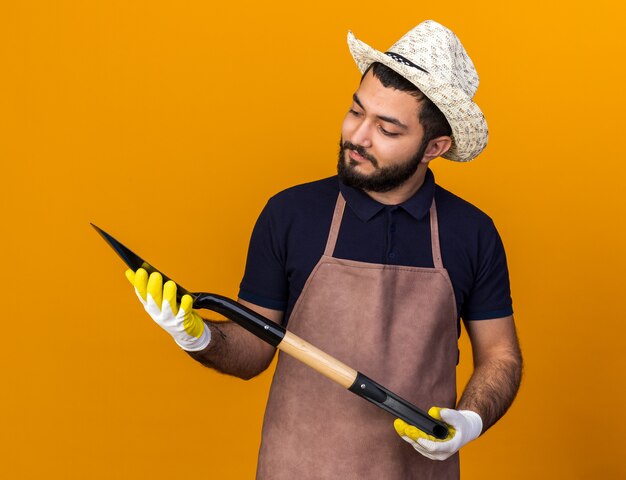 impressed young caucasian male gardener wearing gardening hat and gloves holding and looking at spade isolated on orange wall with copy space