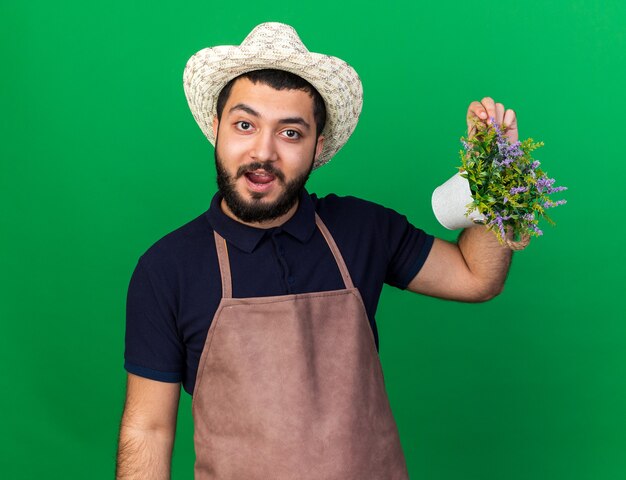 impressed young caucasian male gardener wearing gardening hat holding flowerpot isolated on green wall with copy space