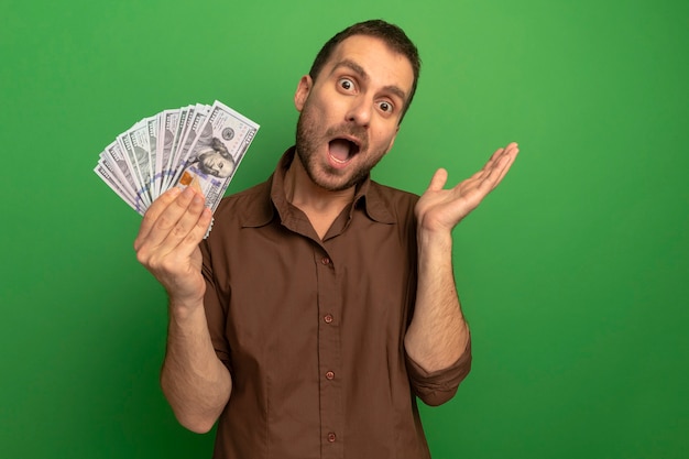 Impressed young caucasian man holding money showing empty hand  isolated on green wall with copy space