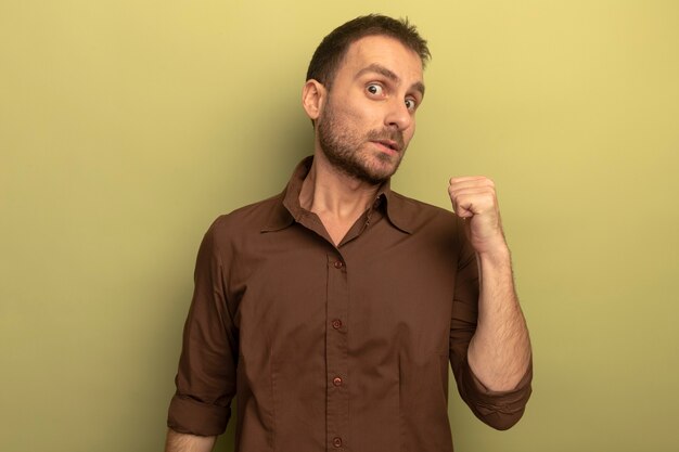 Impressed young caucasian man looking at camera pointing behind isolated on olive green background with copy space