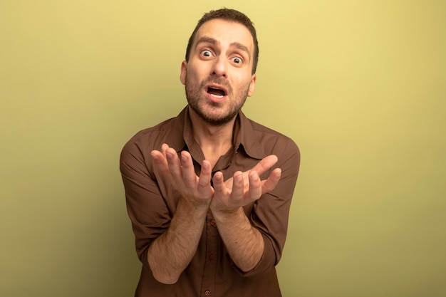 Impressed young caucasian man looking at camera showing empty hands isolated on olive green background with copy space