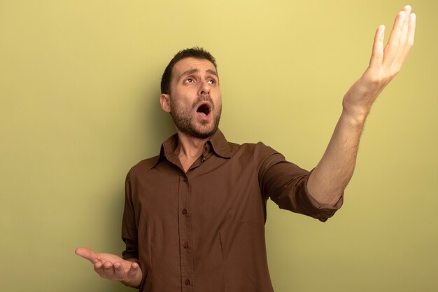 Impressed young caucasian man looking up raising hand up showing empty hand isolated on olive green background