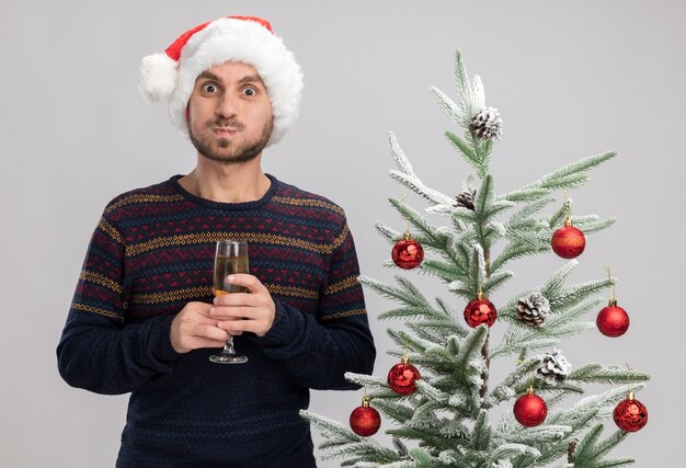 흰색 배경에 고립 된 카메라 퍼핑 뺨을보고 샴페인 잔을 들고 크리스마스 트리 근처 서 크리스마스 모자를 쓰고 감동 젊은 백인 남자