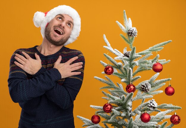 Impressed young caucasian man wearing christmas hat standing near decorated christmas tree keeping hands crossed on arms looking up isolated on orange wall