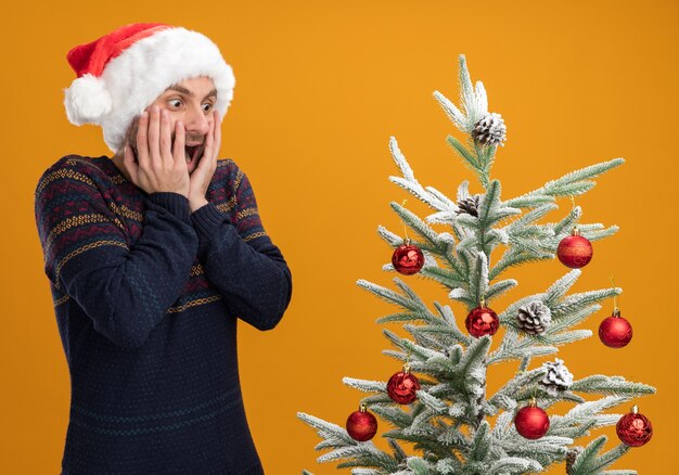 오렌지 배경에 고립 된 나무를보고 얼굴에 손을 유지 장식 된 크리스마스 트리 근처에 서 크리스마스 모자를 쓰고 감동 젊은 백인 남자