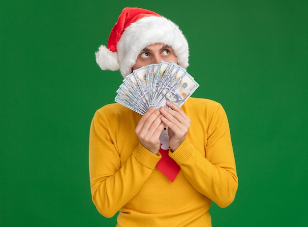 Impressed young caucasian man wearing christmas hat and tie holding money looking up from behind it isolated on green background