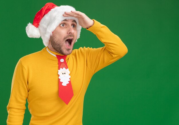 Impressed young caucasian man wearing christmas hat and tie keeping hand on forehead looking at side into distance isolated on green background