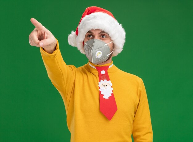 Impressed young caucasian man wearing christmas hat and tie with protective mask looking and pointing at side isolated on green wall