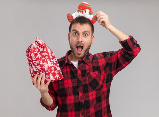 Impressed young caucasian man wearing christmas headband holding christmas sack grabbing headband looking at camera isolated on white background