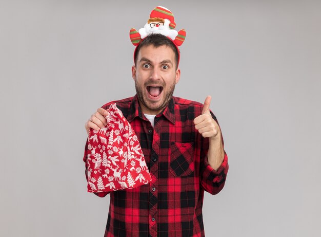 Impressed young caucasian man wearing christmas headband holding christmas sack looking at camera showing thumb up isolated on white background