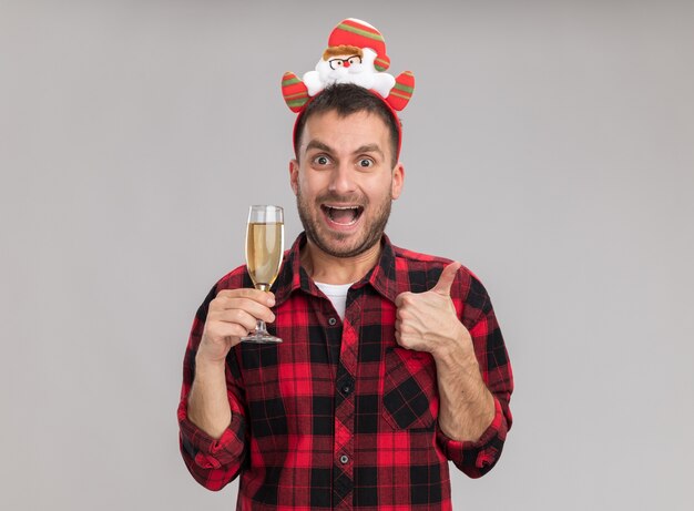 Impressed young caucasian man wearing christmas headband holding glass of champagne  showing thumb up isolated on white wall with copy space