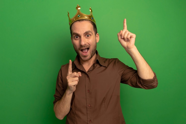 Impressed young caucasian man wearing crown looking at camera pointing up isolated on green background with copy space