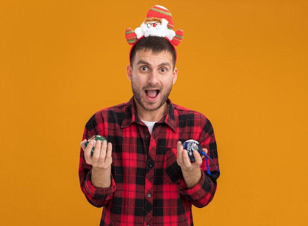 Impressionato giovane uomo caucasico indossando la fascia di babbo natale che tiene le bagattelle di natale isolate sulla parete arancione con lo spazio della copia