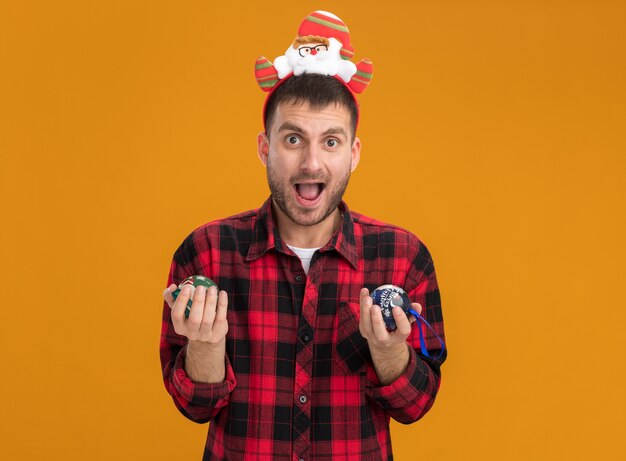 Impressed young caucasian man wearing santa claus headband  holding christmas baubles isolated on orange wall with copy space