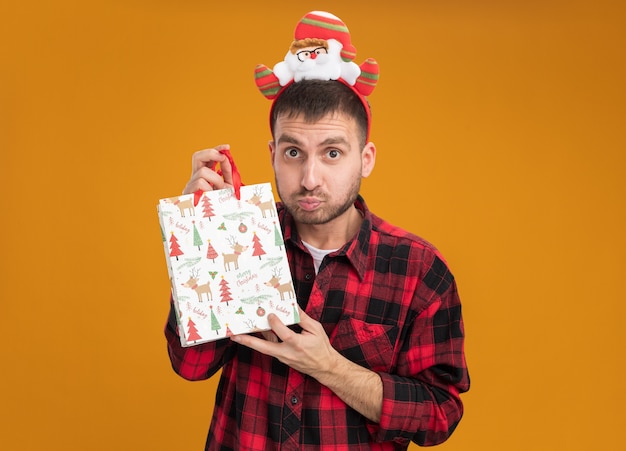 Impressed young caucasian man wearing santa claus headband holding christmas gift bag looking at camera isolated on orange background