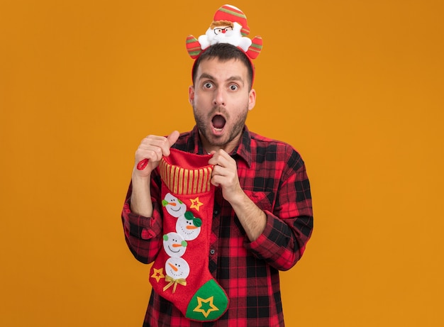 Impressed young caucasian man wearing santa claus headband holding christmas stocking opening it looking at camera isolated on orange background