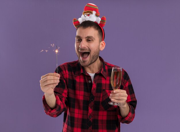 Impressed young caucasian man wearing santa claus headband holding holiday sparkler and glass of champagne looking at sparkler isolated on purple background