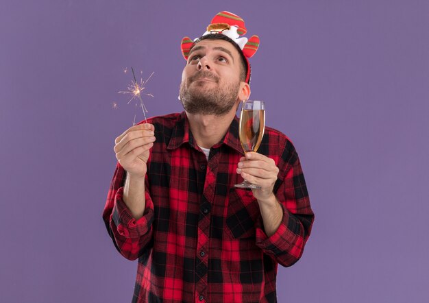 Impressed young caucasian man wearing santa claus headband holding holiday sparkler and glass of champagne looking up isolated on purple wall with copy space