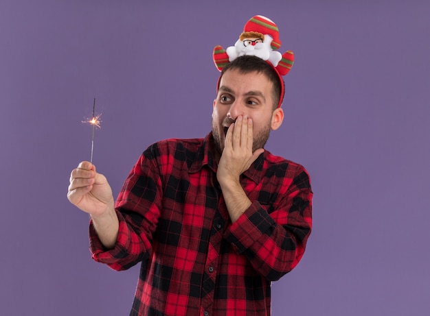 Impressed young caucasian man wearing santa claus headband holding and looking at holiday sparkler keeping hand on face isolated on purple background