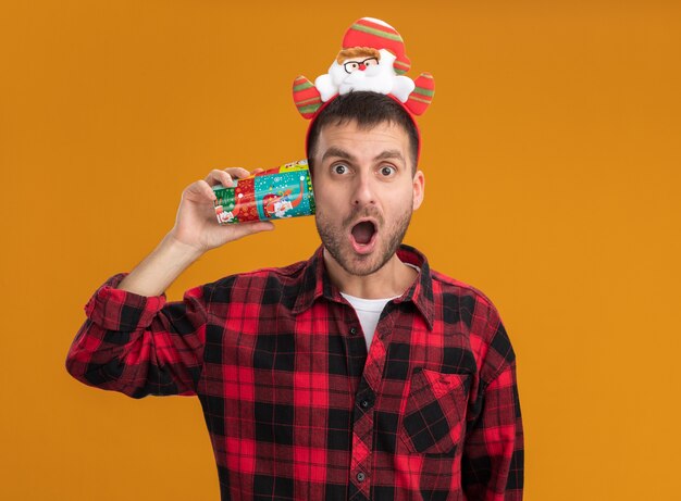 Impressed young caucasian man wearing santa claus headband holding plastic christmas cup next to ear  isolated on orange wall