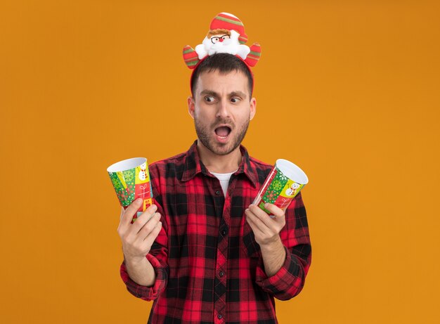 Impressed young caucasian man wearing santa claus headband holding plastic christmas cups looking at one of them isolated on orange background