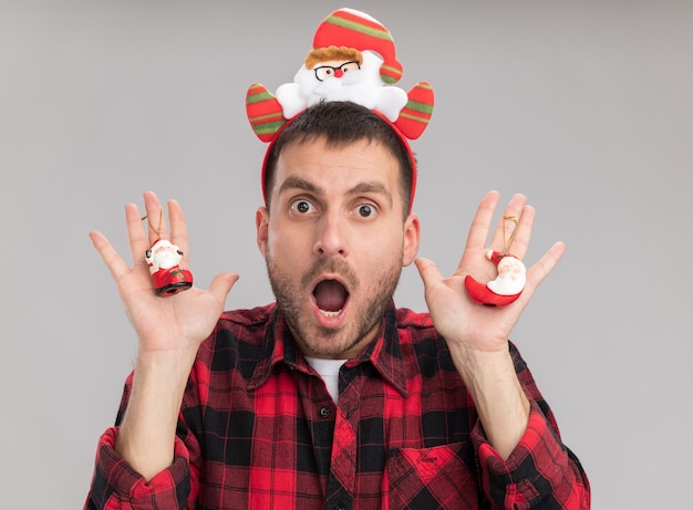 Impressed young caucasian man wearing santa claus headband holding santa claus christmas ornaments  isolated on white wall