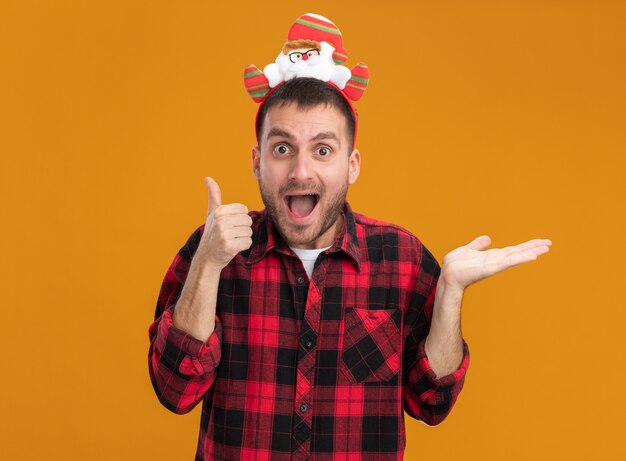Impressed young caucasian man wearing santa claus headband looking at camera showing thumb up and empty hand isolated on orange background