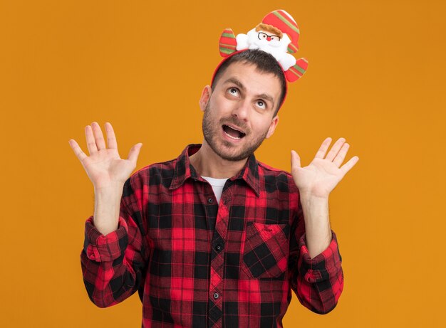 Impressed young caucasian man wearing santa claus headband showing empty hands looking up isolated on orange background