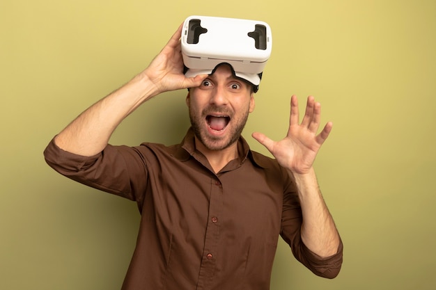 복사 공간 올리브 녹색 배경에 고립 된 빈 손을 보여주는 카메라를보고 그것을 잡는 이마에 VR 헤드셋을 착용하는 감동 된 젊은 백인 남자