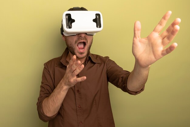 올리브 녹색 배경에 고립 된 손을 뻗어 측면을보고 VR 헤드셋을 착용하는 감동 된 젊은 백인 남자