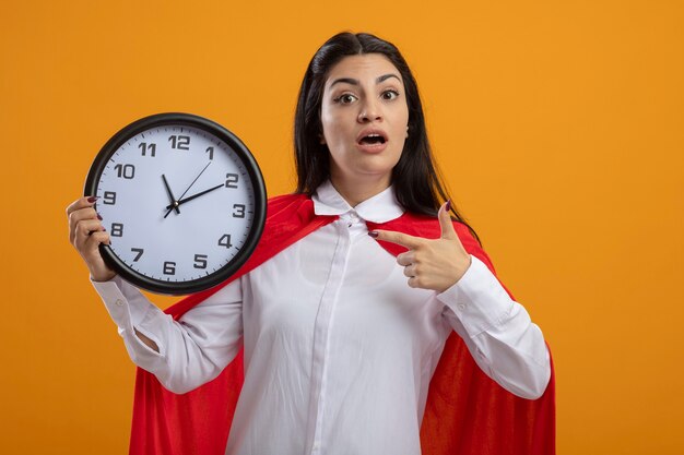 Impressed young caucasian superhero girl holding and pointing at clock looking at camera isolated on orange background