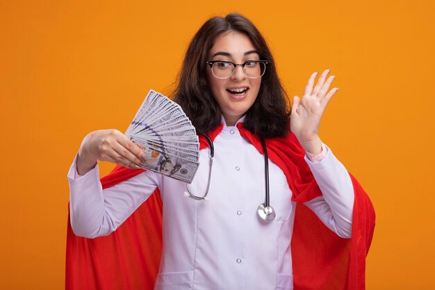 Impressed young caucasian superhero girl wearing doctor uniform and stethoscope with glasses holding and looking at money showing empty hand isolated on wall