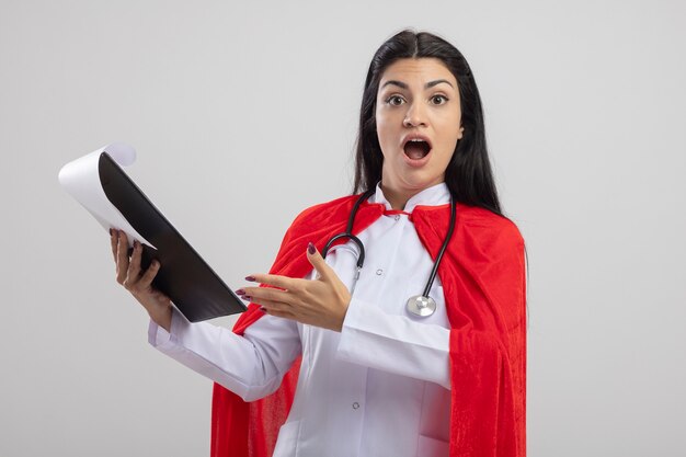 Impressed young caucasian superhero girl wearing stethoscope holding clipboard looking at camera isolated on white background with copy space