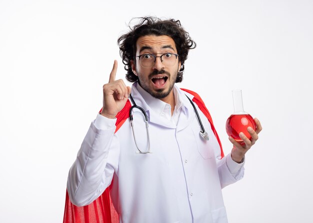 Impressed young caucasian superhero man in optical glasses wearing doctor uniform with red cloak and with stethoscope around neck holds red chemical liquid in glass flask and points up