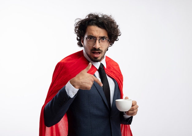 Impressed young caucasian superhero man in optical glasses wearing suit with red cloak holds and points at cup 
