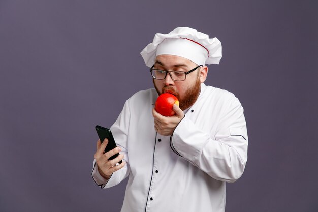 보라색 배경에 격리된 사과를 먹으면서 안경을 쓰고 모자를 들고 휴대전화를 쳐다보는 젊은 셰프