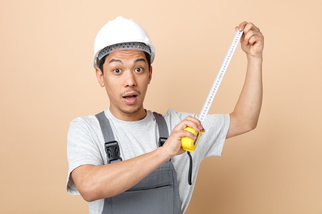 Impressed young construction worker wearing safety helmet and uniform holding tape meter 