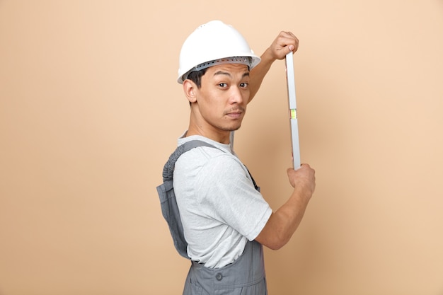 Impressed young construction worker wearing safety helmet and uniform standing in profile view holding level ruler