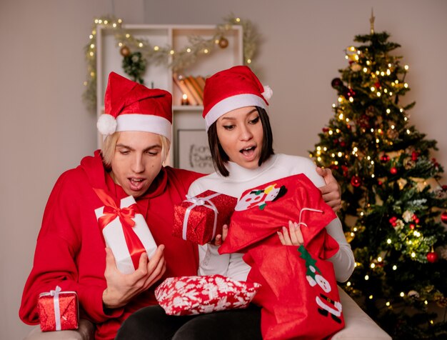 Impressed young couple at home at christmas time wearing santa hat sitting on armchair holding and looking at christmas gift packages and sacks in living room