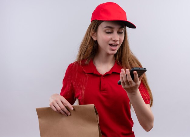 Impressed young delivery girl in red uniform holding and looking at mobile phone on isolated white space