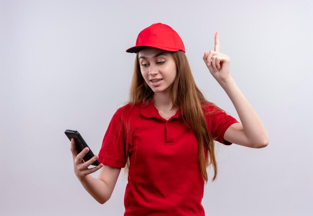 Impressed young delivery girl in red uniform holding and looking at mobile phone with raised finger on isolated white space