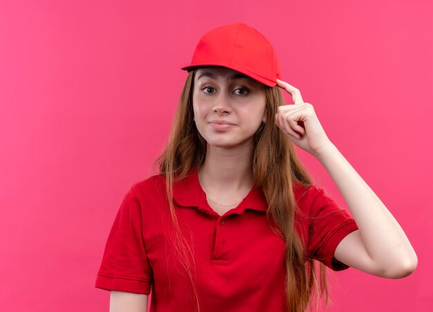 Impressed young delivery girl in red uniform putting finger on head on isolated pink space with copy space