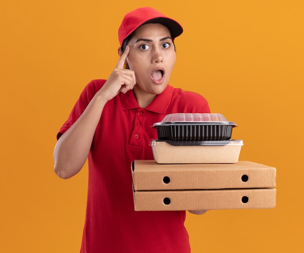 Impressed young delivery girl wearing uniform and cap holding pizza boxes with food containers putting finger on temple isolated on orange wall