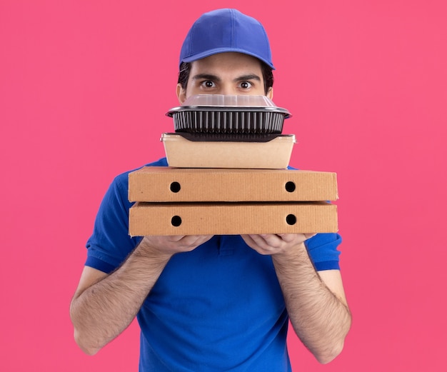 Impressionato giovane fattorino in uniforme blu e cappuccio che tiene i pacchetti di pizza con contenitore per alimenti e pacchetto di carta alimentare su di loro guardando davanti da dietro di loro isolato sul muro rosa