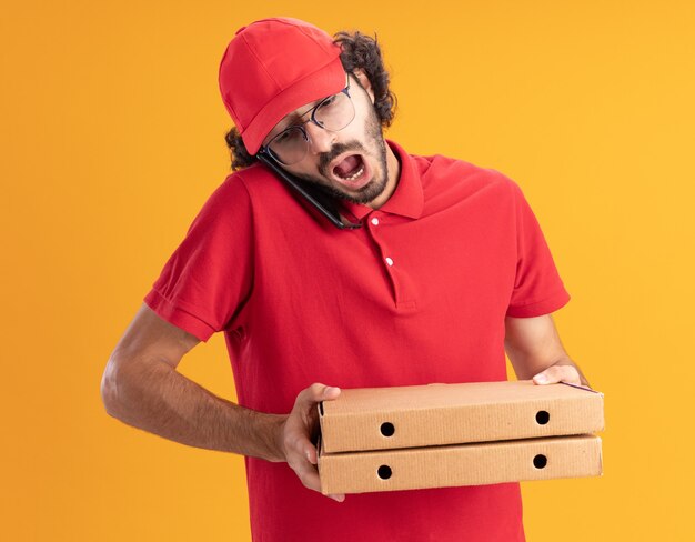 Impressionato giovane fattorino in uniforme rossa e berretto con gli occhiali che tengono i pacchetti di pizza parlando al telefono guardando in basso isolato sul muro arancione