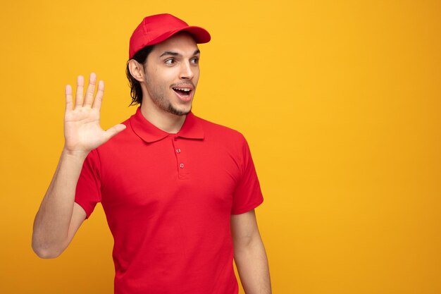 impressed young delivery man wearing uniform and cap loking at side showing hi gesture isolated on yellow background with copy space