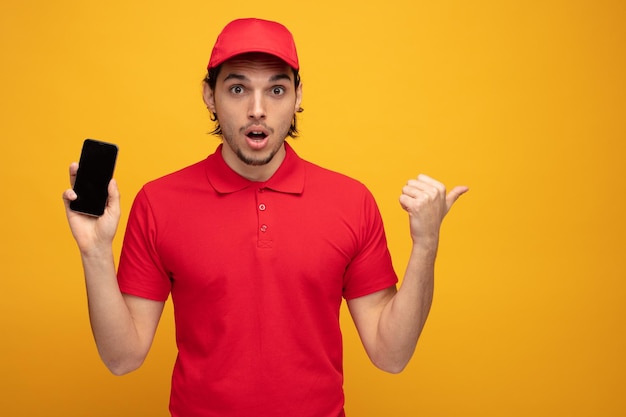 Impressionato giovane fattorino che indossa uniforme e cappuccio guardando la fotocamera che mostra il telefono cellulare che punta al lato isolato su sfondo giallo